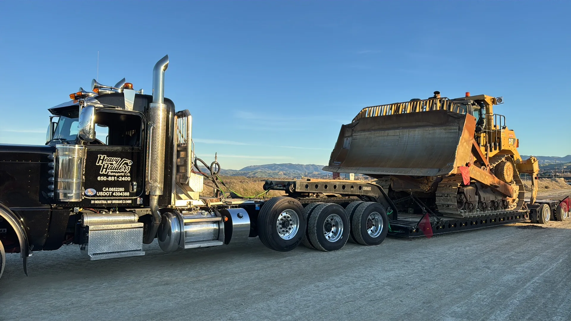 Heavy Haulers lowboy trailer transporting a massive CAT D-series bulldozer at golden hour in the California desert